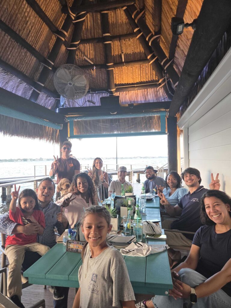 Group photo of all attendees at the SoFlo SubTo Monthly Mixer Treasure Coast event, smiling and throwing peace signs under the tiki pavilion with waterfront in background, including families with kids and a dog