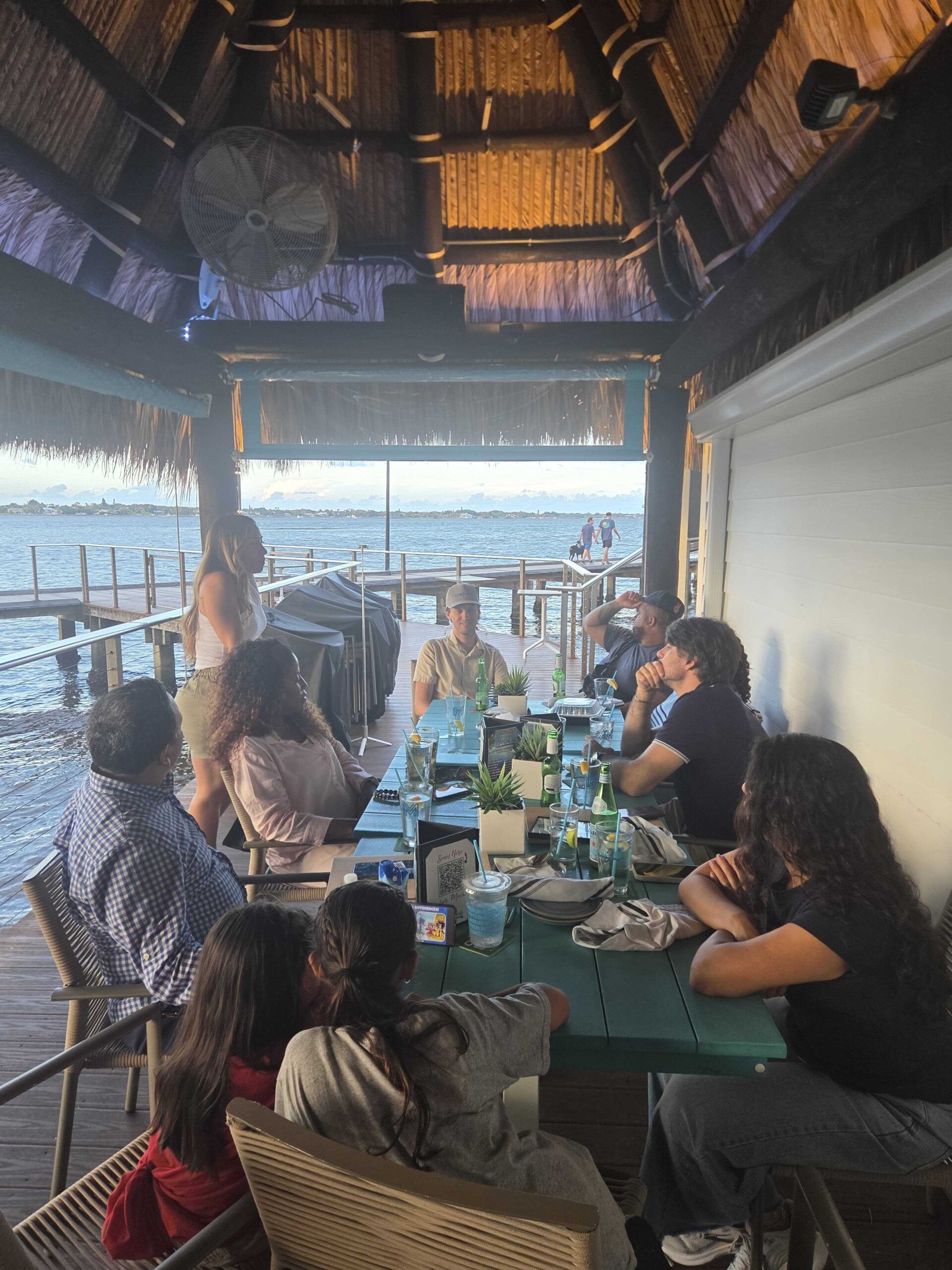 Group of real estate investors networking at a long table under a tiki-style waterfront pavilion at sunset with drinks and the water visible in the background